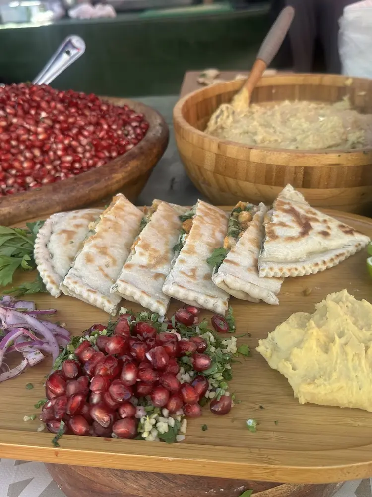 Wooden board with folded gözleme wedges, sumac red onion, pomegranate and bulgur salad, hummus and lime